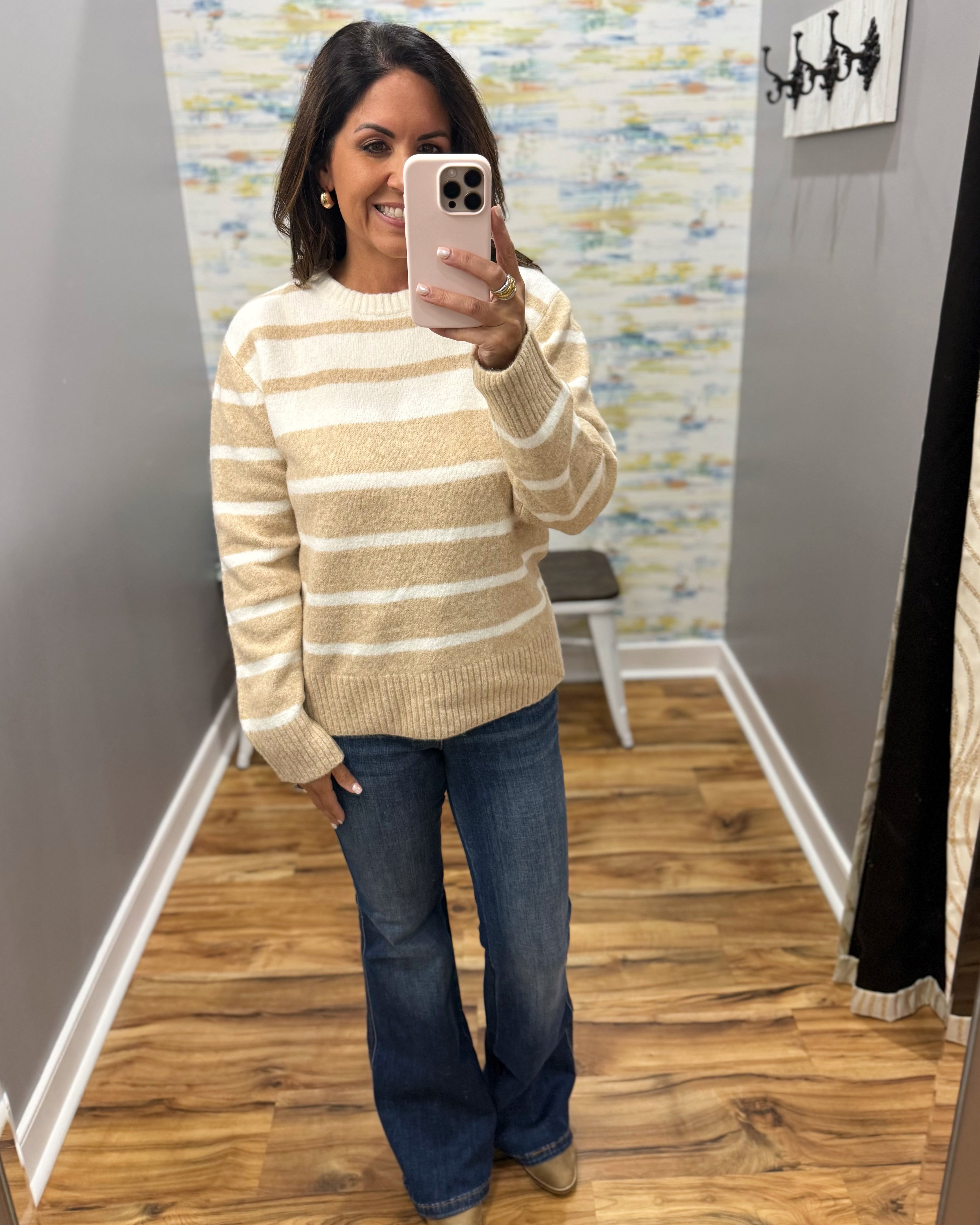 A woman takes a mirror selfie in a fitting room wearing a beige and white striped knit sweater, blue jeans, and tan boots.