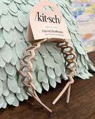 A set of three Kit'sch zigzag headbands in dusty pink, beige, and taupe, attached to a paper hanger card and displayed on a wooden surface in front of a mint green feather pillow.