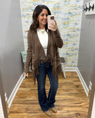 A woman takes a mirror selfie in a fitting room wearing a brown open-front fringed cardigan over a white top, blue jeans, and tan boots.