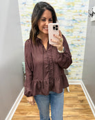 A woman takes a mirror selfie wearing a long-sleeve, button-front blouse with a ruffled front and peplum hem in a dark brown color with a small white floral pattern, paired with blue jeans.