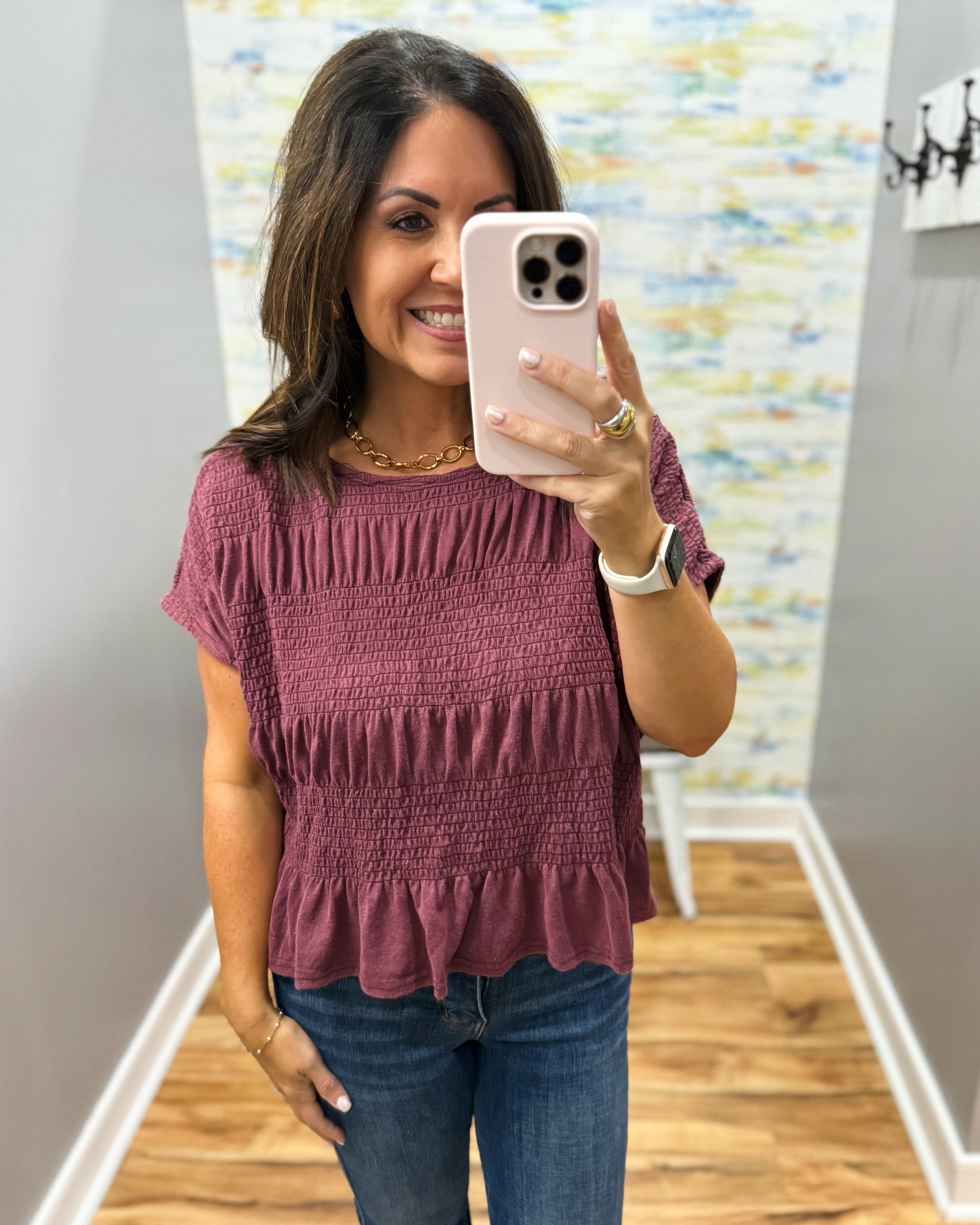 A woman taking a mirror selfie wearing a dusty rose short-sleeve smocked blouse and blue jeans in a hallway.