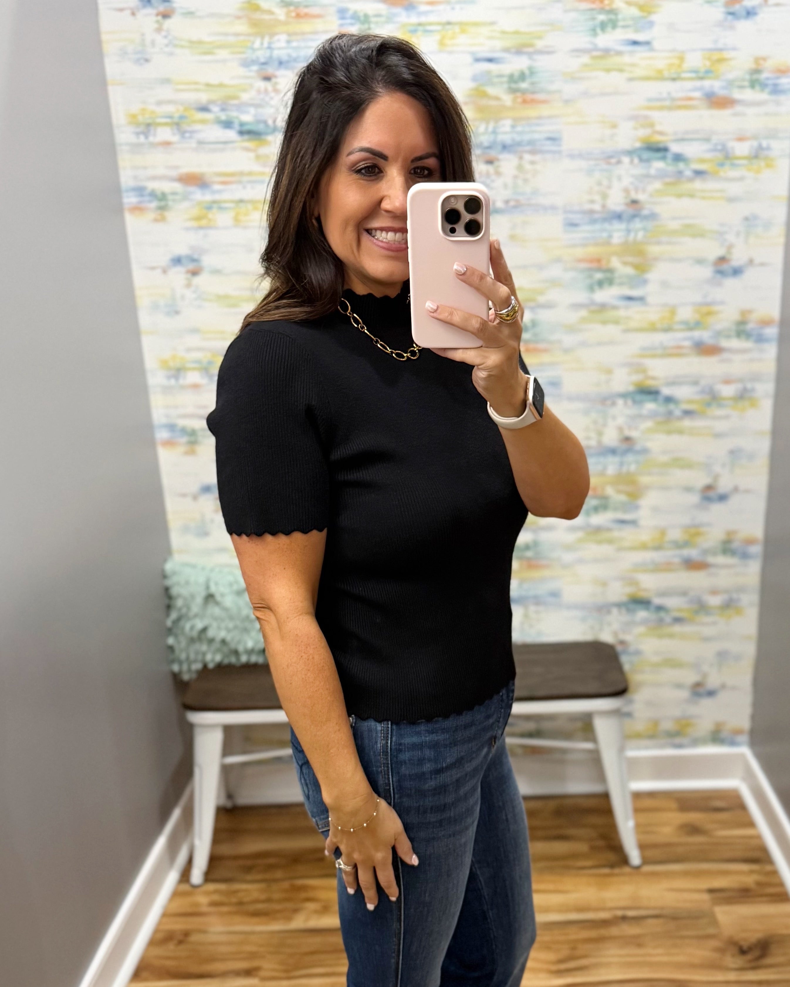 A woman taking a mirror selfie in a boutique, wearing a black short-sleeve mock neck top with scalloped edges and blue jeans.