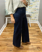 A person wearing dark navy wide-leg jeans, a light green and white striped blouse, and brown wedge shoes, standing in a room with wood floors and a bench.