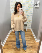A woman takes a mirror selfie in a fitting room wearing a beige knit sweater with white contrast trim, blue flare jeans, and metallic gold shoes.