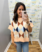 A woman takes a mirror selfie wearing a short-sleeve knit sweater with a multi-colored argyle pattern in peach, beige, brown, navy, and light blue, paired with blue jeans.