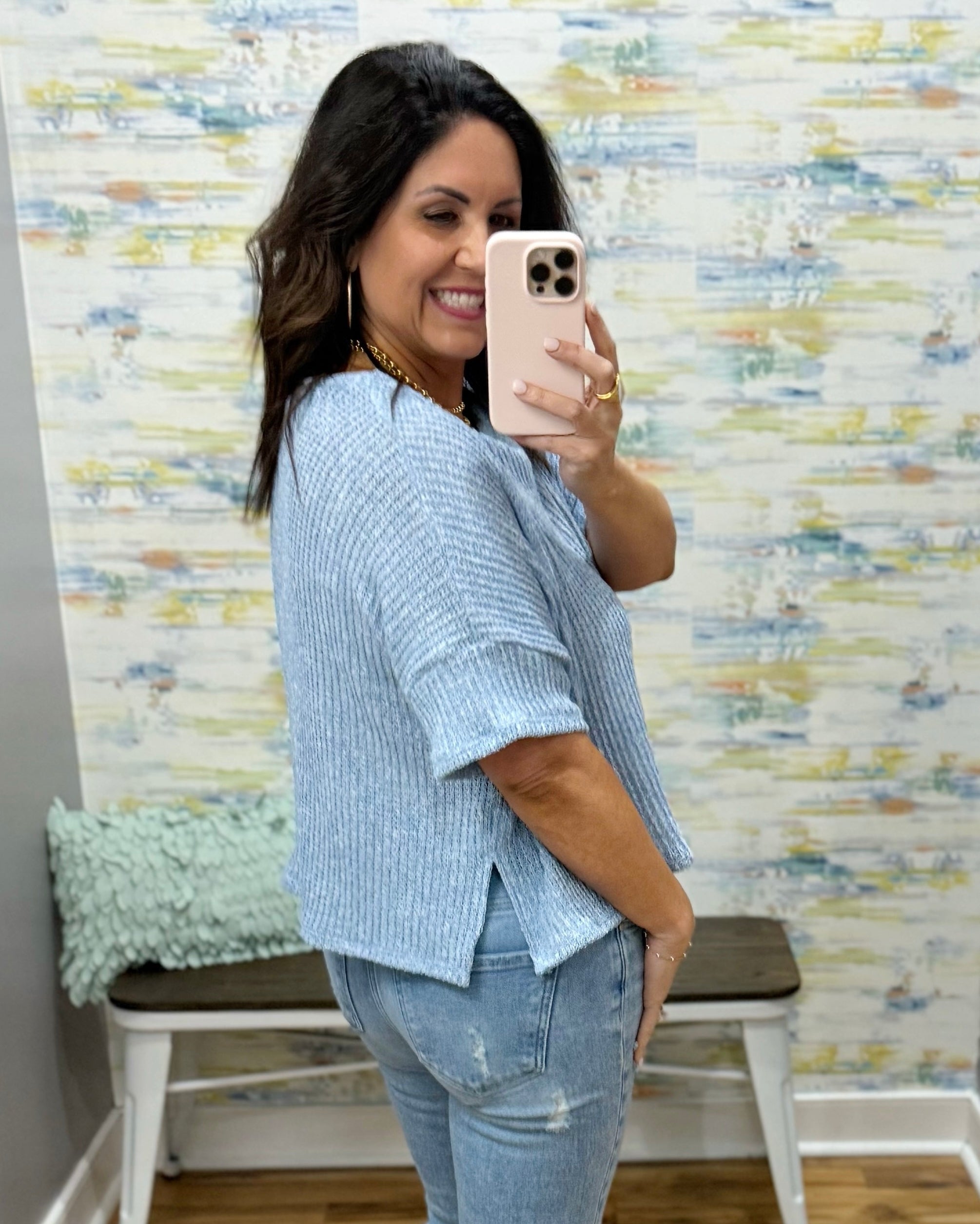 Woman taking a mirror selfie wearing a light blue shirt and jeans in front of a textured wall.