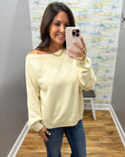 A woman takes a mirror selfie wearing a pale cream off-the-shoulder knit sweater and blue jeans in a fitting room with wood floors and a patterned wall.
