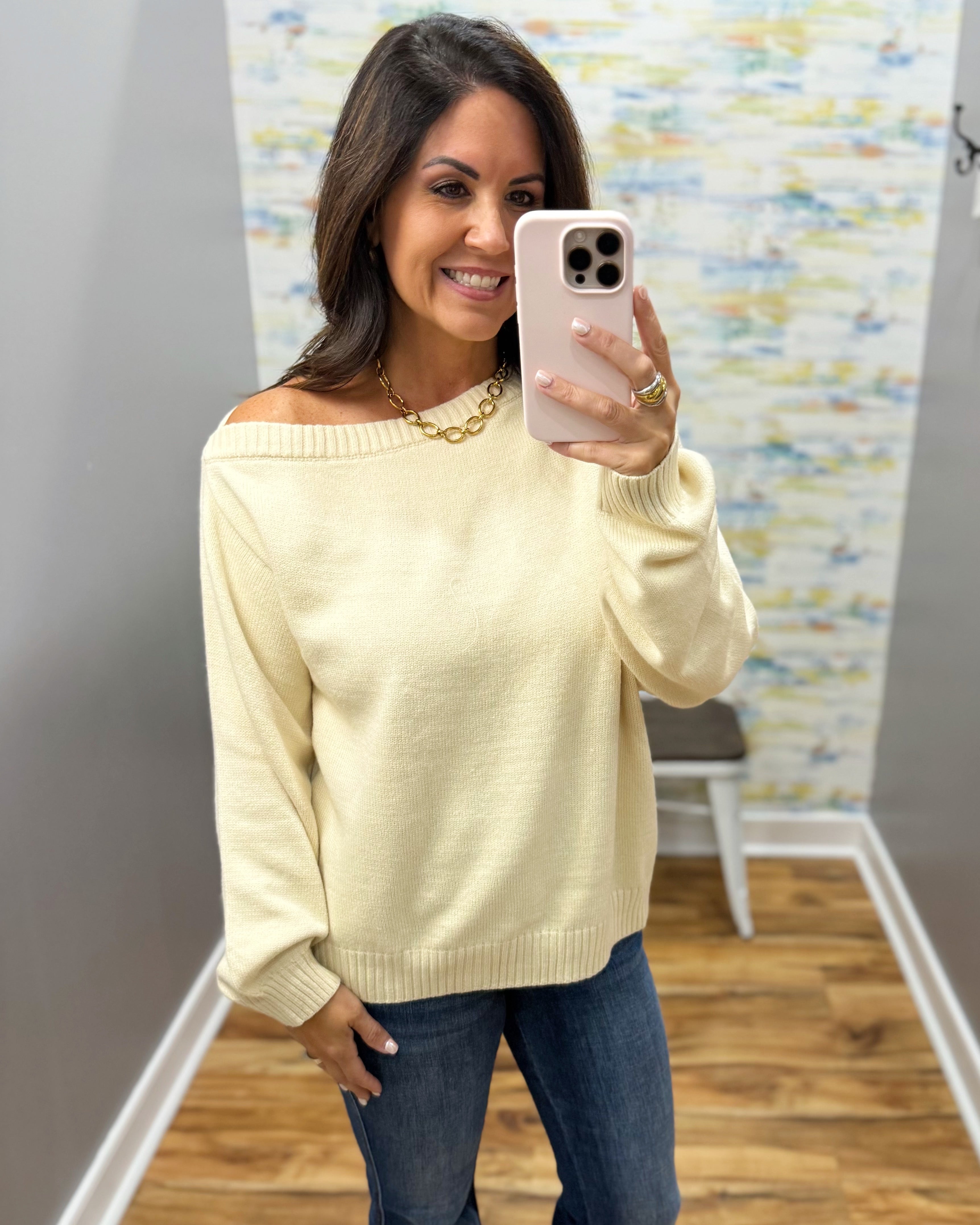 A woman takes a mirror selfie wearing a pale cream off-the-shoulder knit sweater and blue jeans in a fitting room with wood floors and a patterned wall.