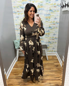 A woman takes a mirror selfie in a fitting room wearing a dark brown long-sleeve maxi dress with a beige floral pattern and a matching waist belt.