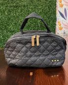 A black quilted nylon cosmetic bag with a top handle, gold-tone zipper pulls, and three gold studs on the front, resting on a wooden table against a green artificial hedge background.