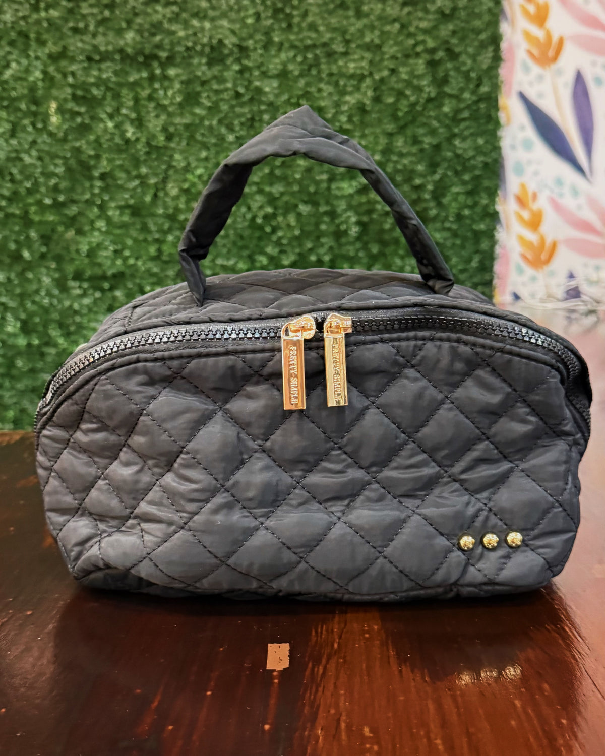 A black quilted nylon cosmetic bag with a top handle, gold-tone zipper pulls, and three gold studs on the front, resting on a wooden table against a green artificial hedge background.