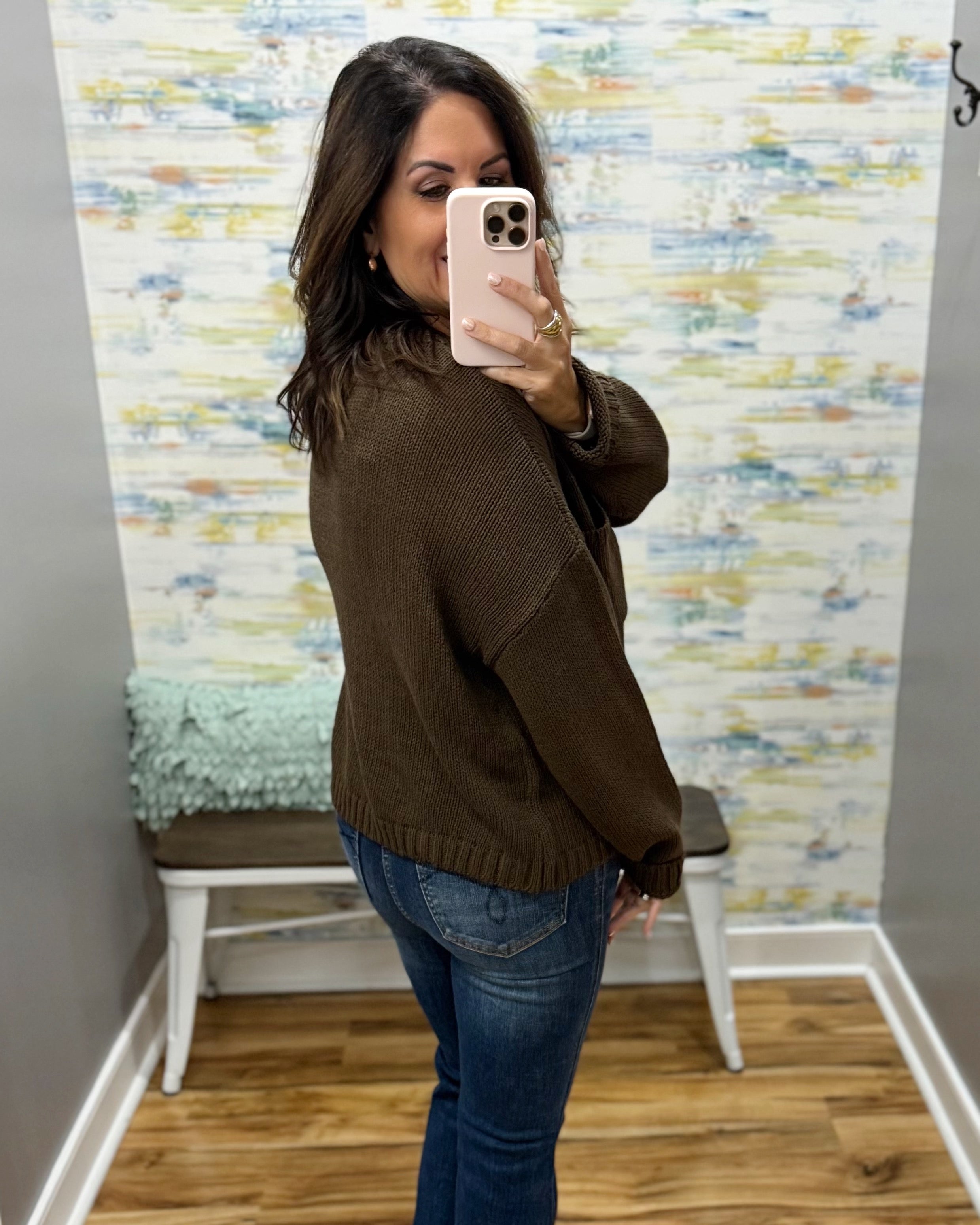 A woman takes a mirror selfie wearing a dark brown knit sweater and blue jeans in a room with abstract wallpaper and a small bench.