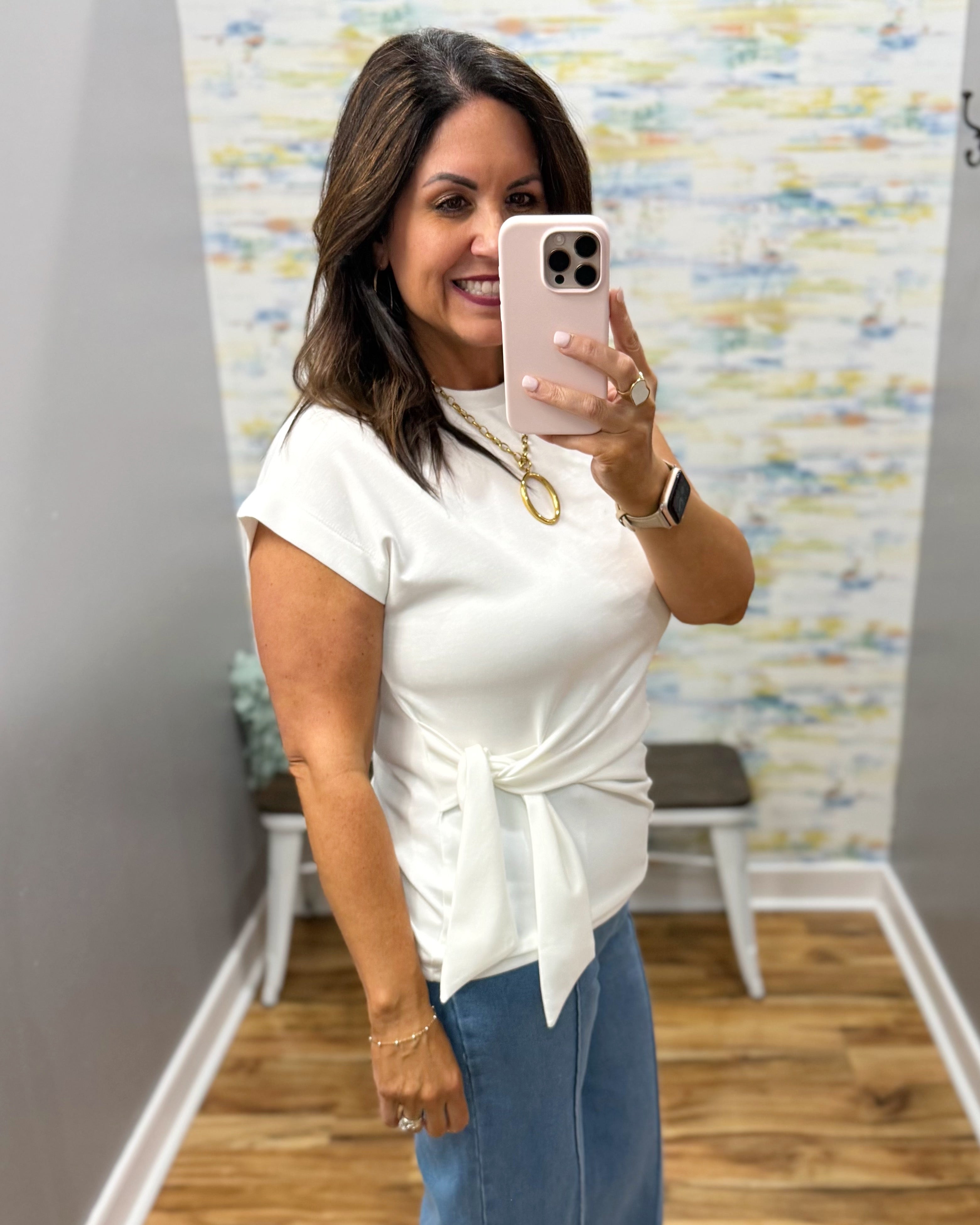Woman taking a mirror selfie wearing a white top and blue jeans in a room with a colorful wall.