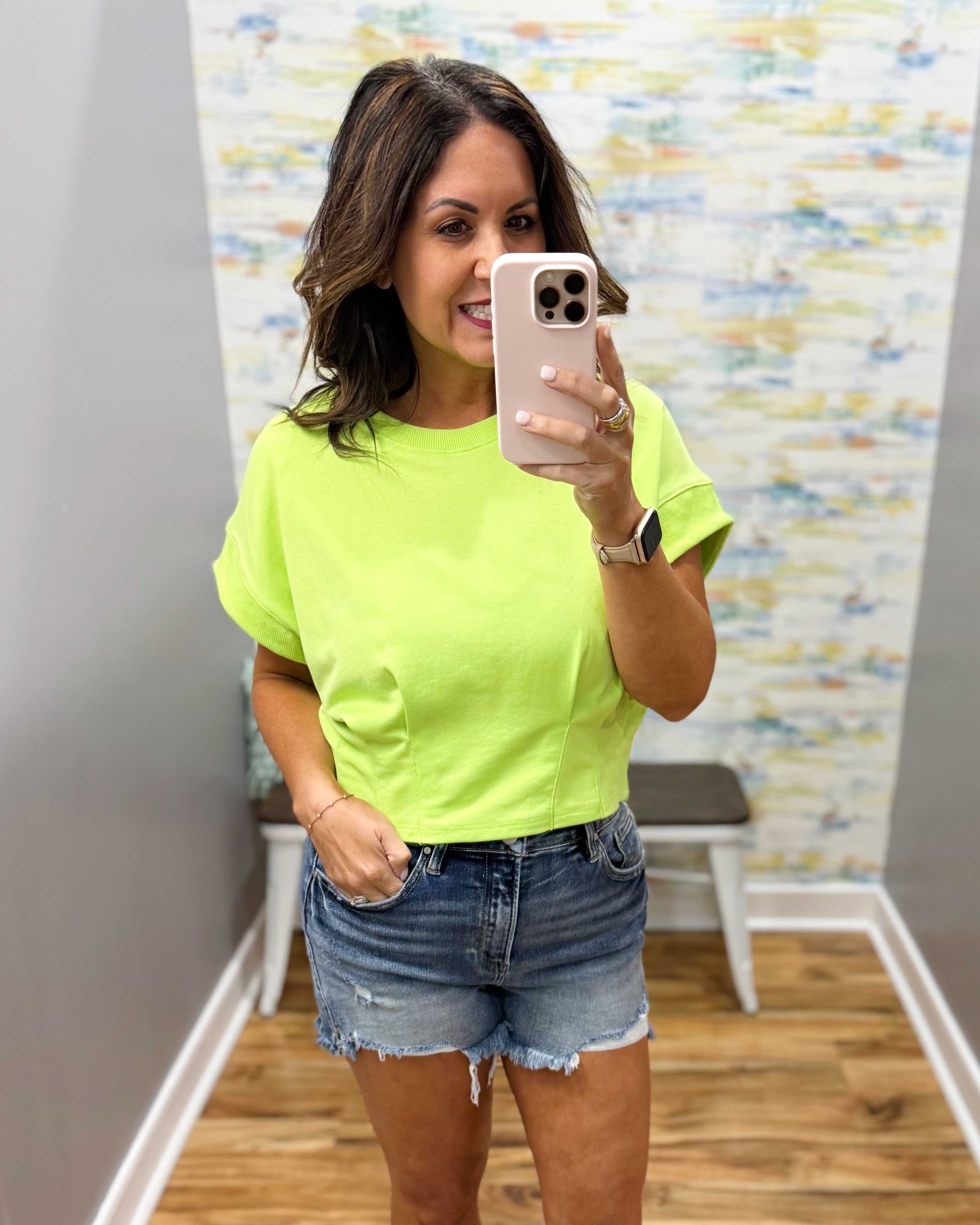Woman taking a mirror selfie wearing a bright green t-shirt and denim shorts in a room with colorful wall art.