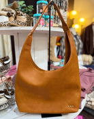 A tan leather hobo shoulder bag with a slouchy shape and a single shoulder strap, displayed on a stand inside a boutique setting.