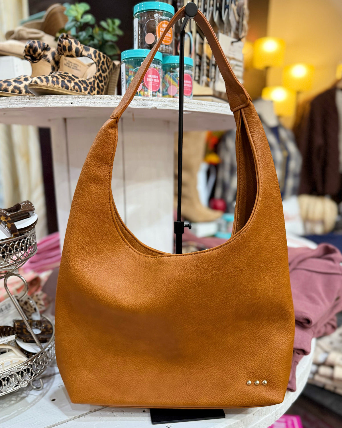 A tan leather hobo shoulder bag with a slouchy shape and a single shoulder strap, displayed on a stand inside a boutique setting.