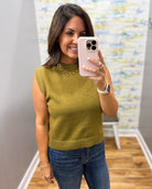 A woman taking a mirror selfie wearing a sleeveless olive green knit sweater vest with blue jeans and a gold chain necklace.