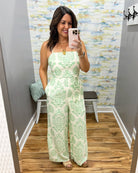 Woman taking a mirror selfie wearing a green and white patterned jumpsuit in a fitting room.
