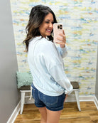 Woman taking a mirror selfie wearing a light blue sweatshirt and denim shorts in a room with colorful wall paper.