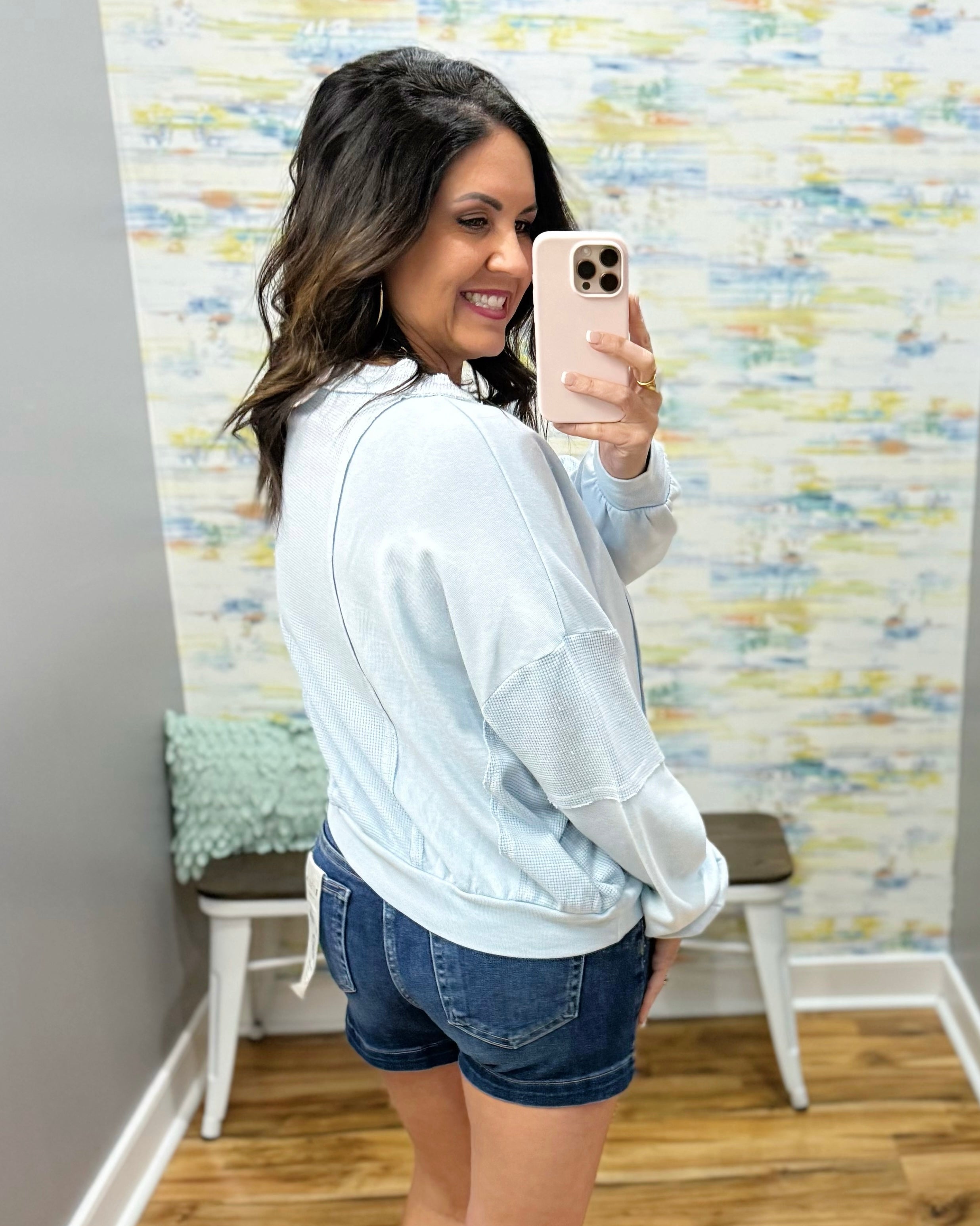 Woman taking a mirror selfie wearing a light blue sweatshirt and denim shorts in a room with colorful wall paper.