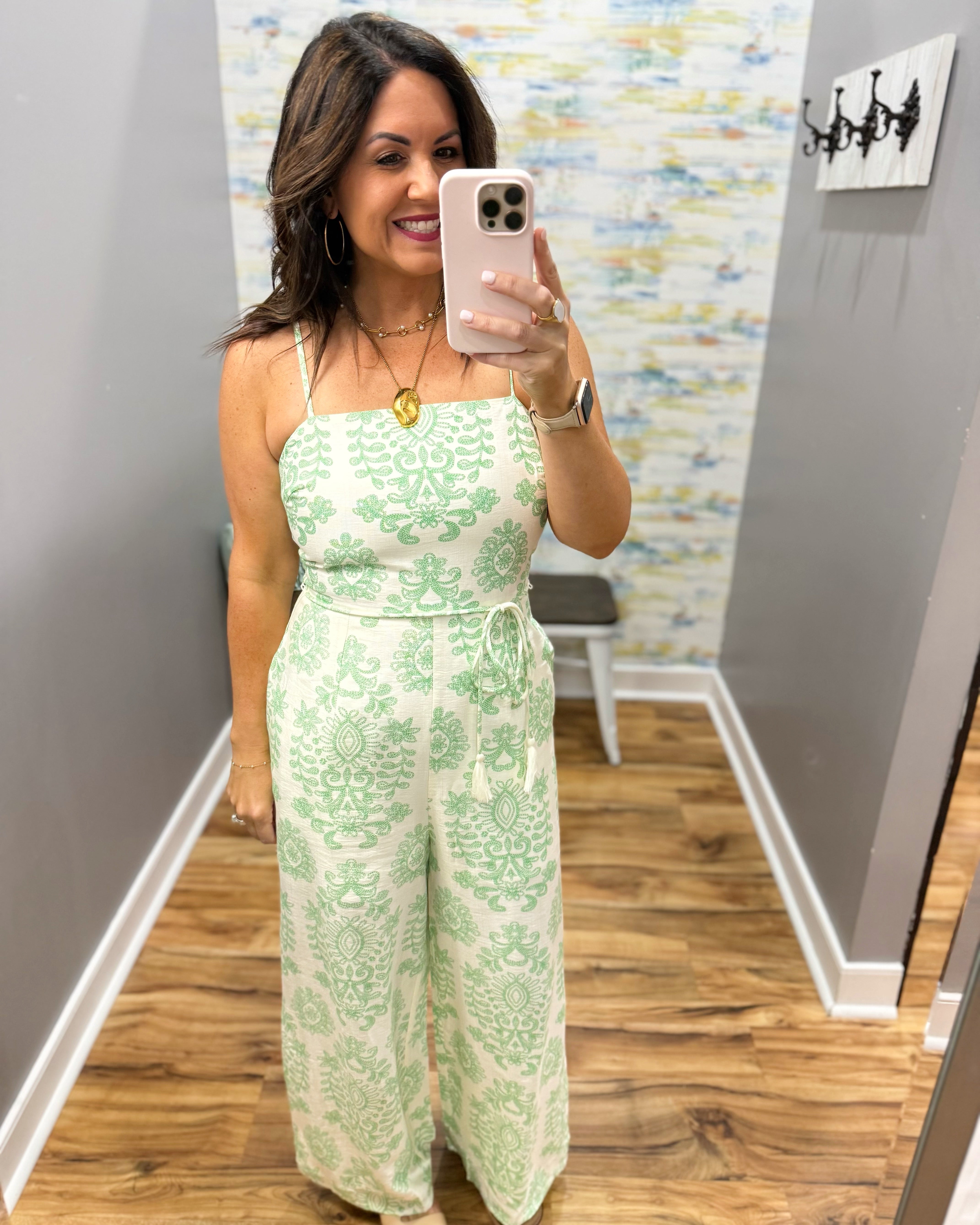 Woman taking a mirror selfie wearing a green floral jumpsuit in a room with a patterned wall.