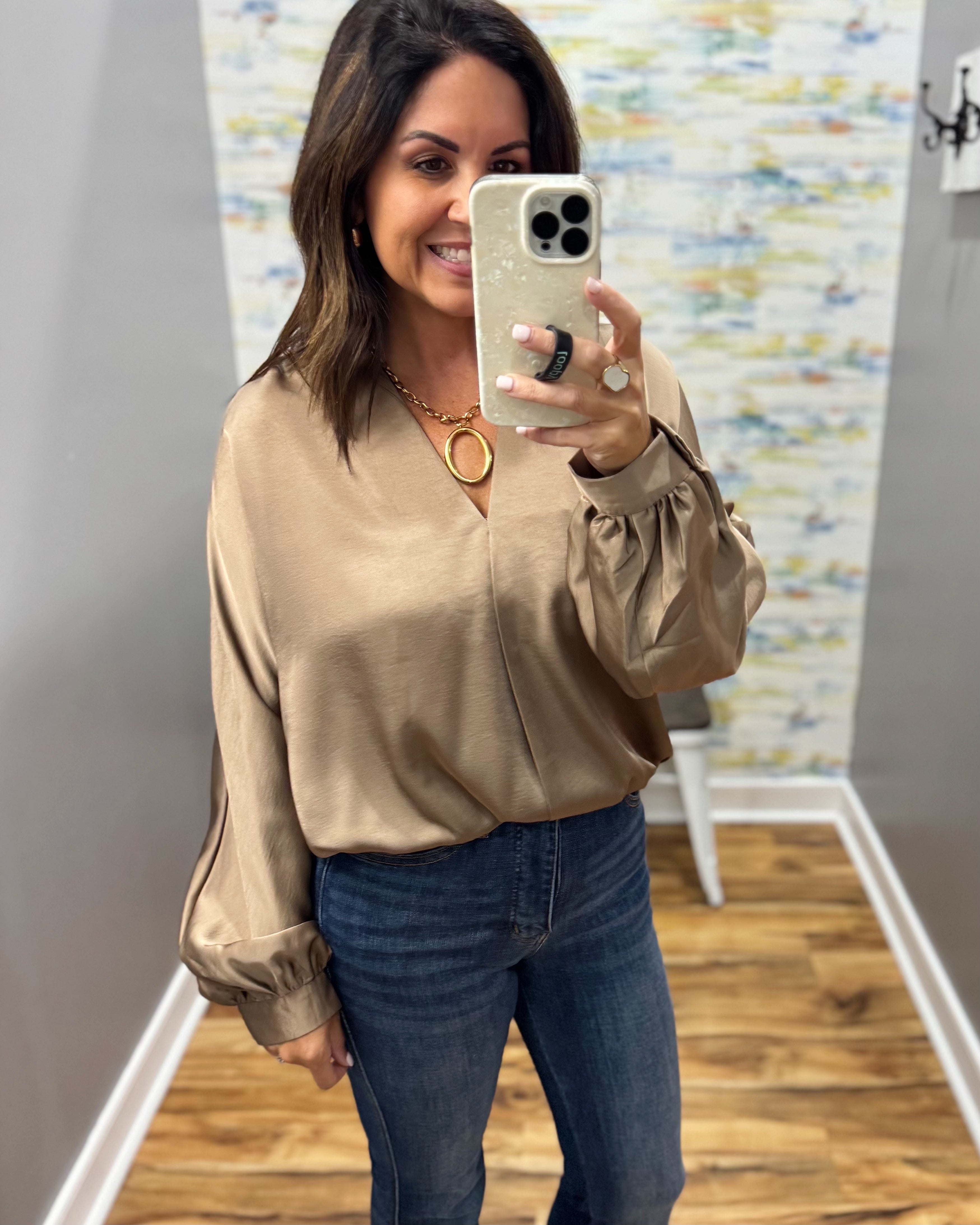 A woman takes a mirror selfie in a fitting room wearing a champagne beige satin blouse with puffed sleeves and blue jeans.