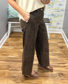 A person wearing brown leopard-print wide-leg pants with a cream top and brown wedge sandals, standing on a wood floor in an indoor room.