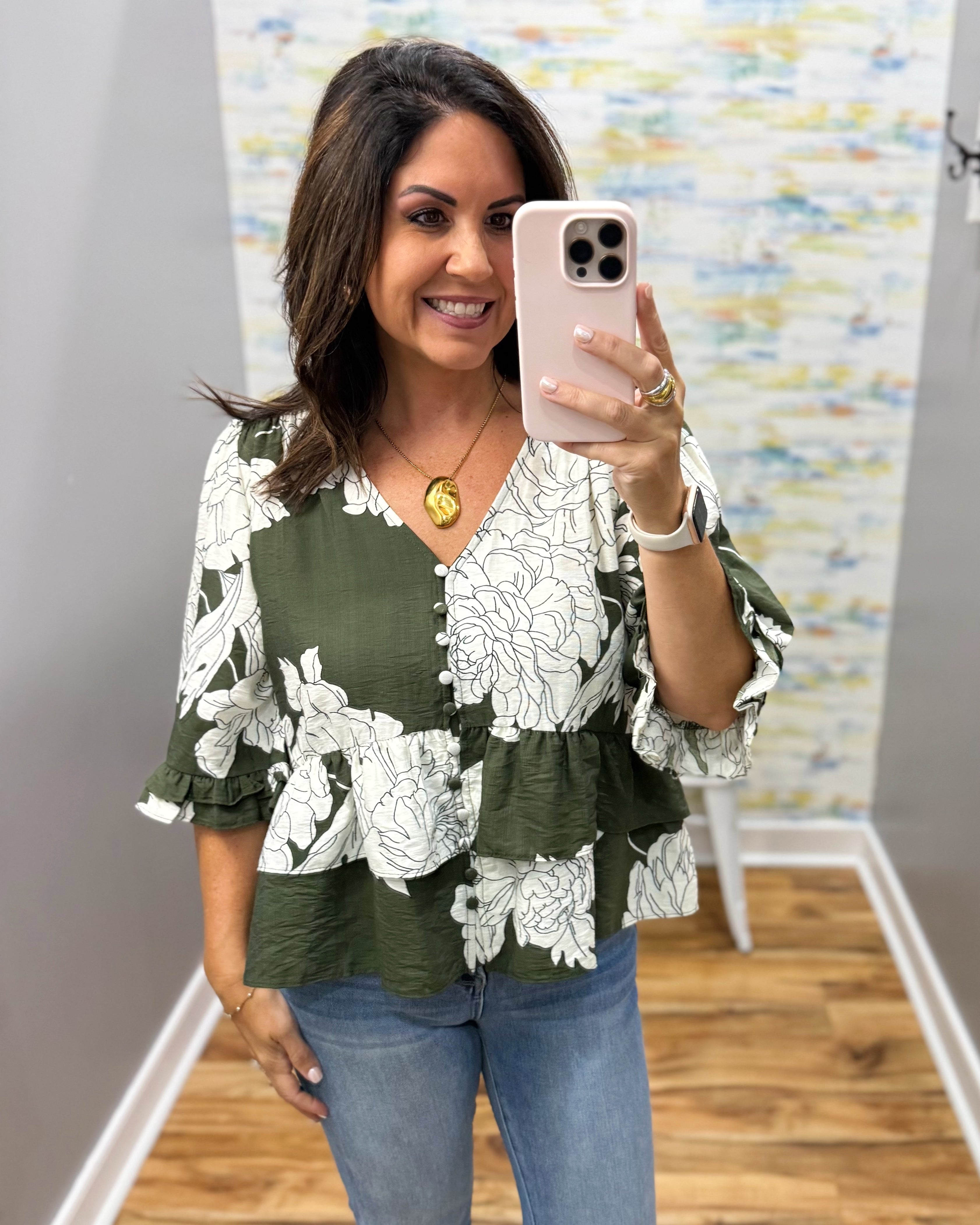 A woman takes a mirror selfie wearing a green and white floral print blouse with tiered sleeves and blue jeans in what appears to be a boutique fitting room.