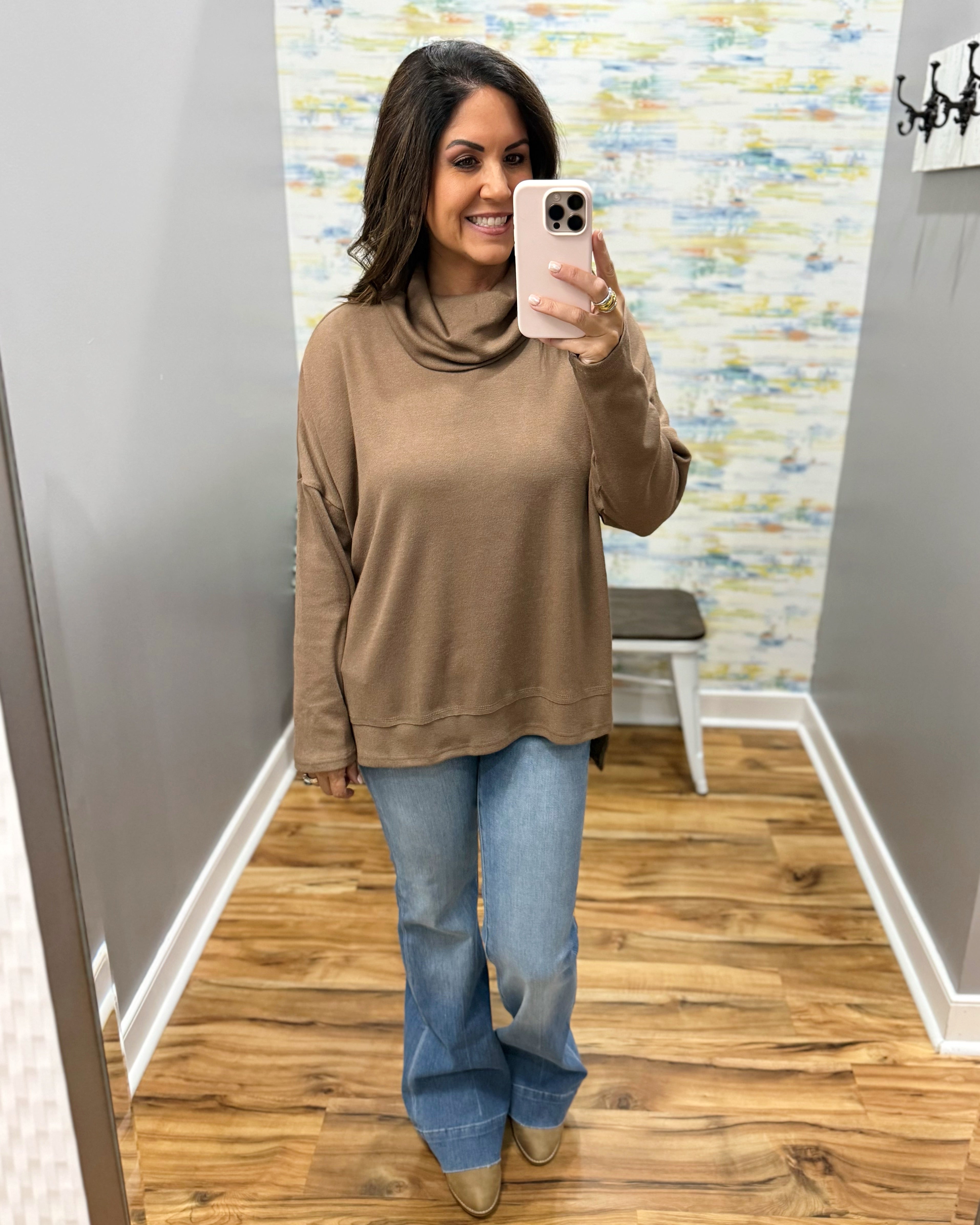A woman takes a mirror selfie in a fitting room wearing a brown cowl-neck knit top and blue flare jeans.