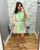 Woman taking a mirror selfie wearing a colorful floral dress in a room with patterned wallpaper.