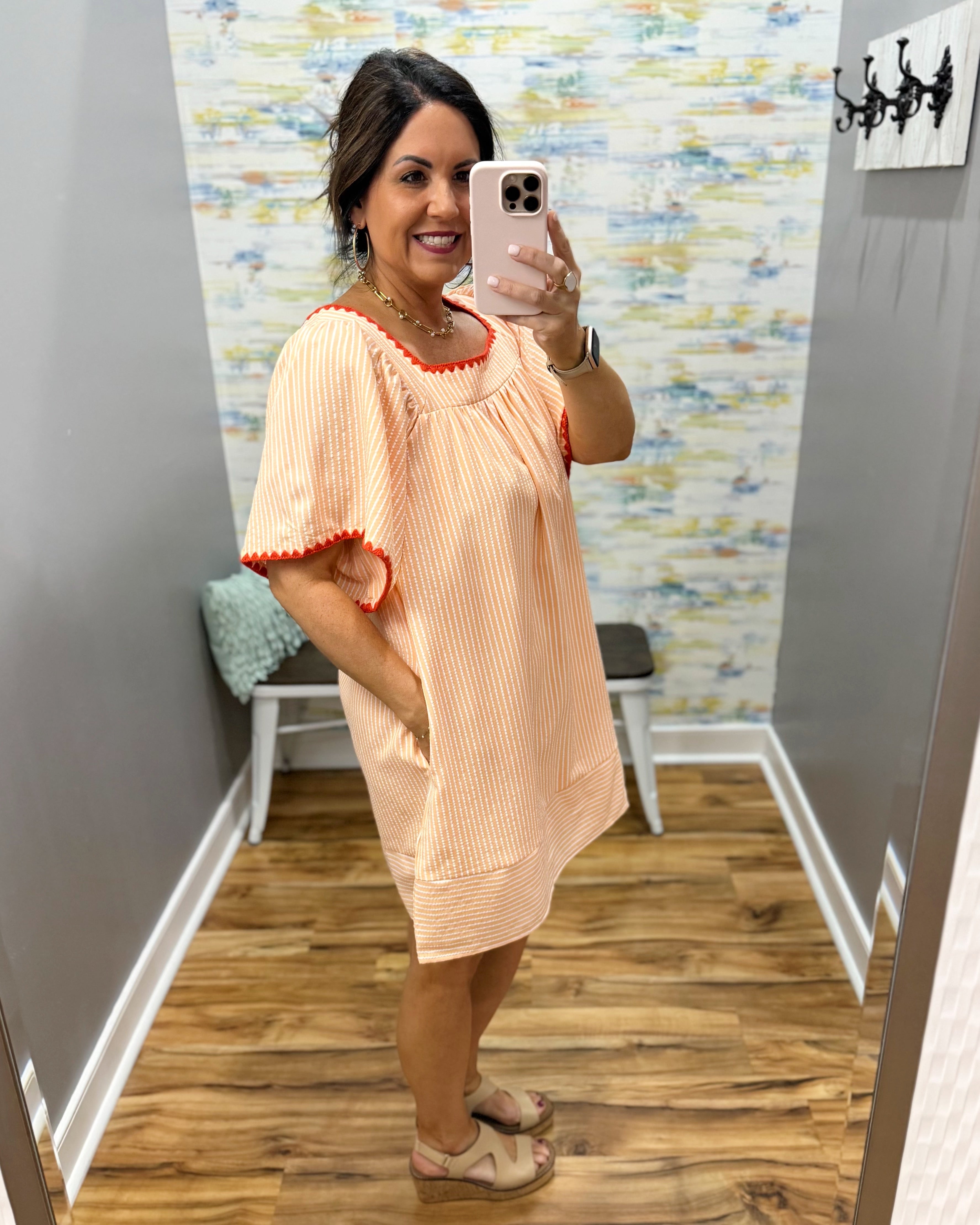 Woman taking a mirror selfie wearing a peach dress with red trim in a room with a colorful wall.