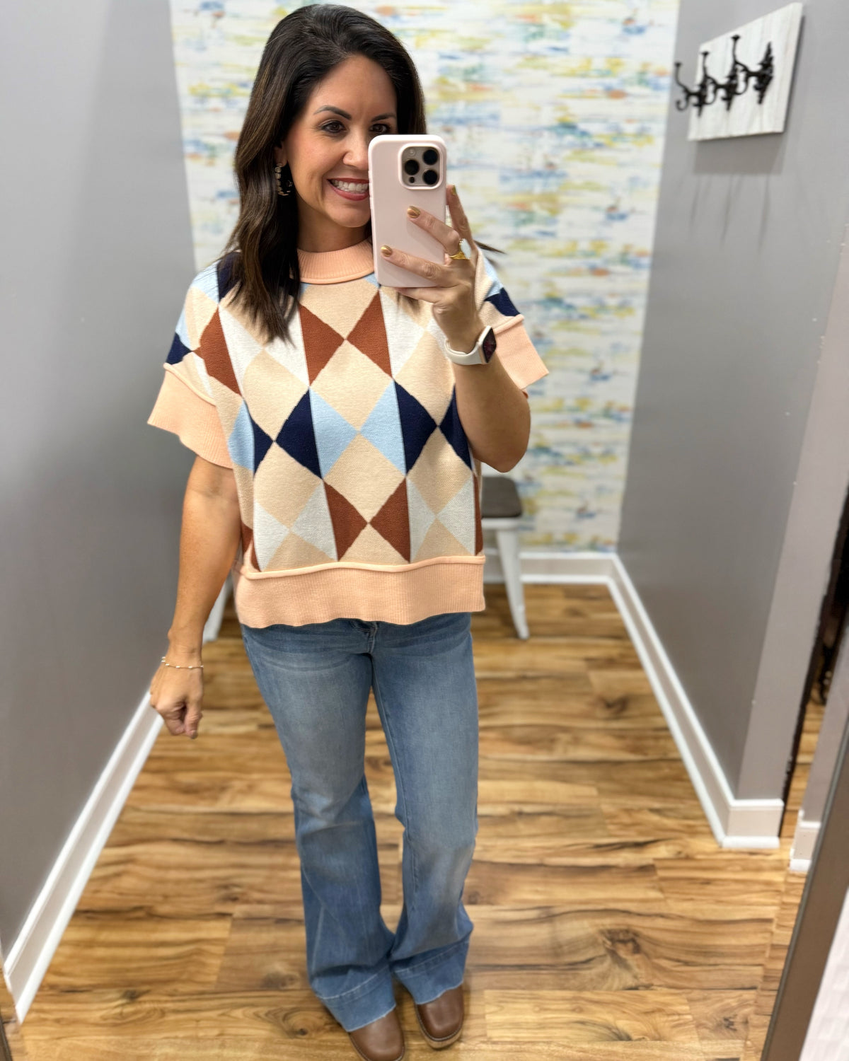 A woman takes a mirror selfie wearing a short-sleeve knit sweater with a geometric diamond pattern in peach, brown, light blue, and navy, paired with blue jeans and brown shoes, in an indoor room with wood floors and gray walls.