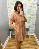 A woman takes a mirror selfie in a fitting room wearing a short-sleeve, collared, tiered shift dress in a warm tan or camel color. The background features a patterned wallpaper, a wooden floor, and a white bench.