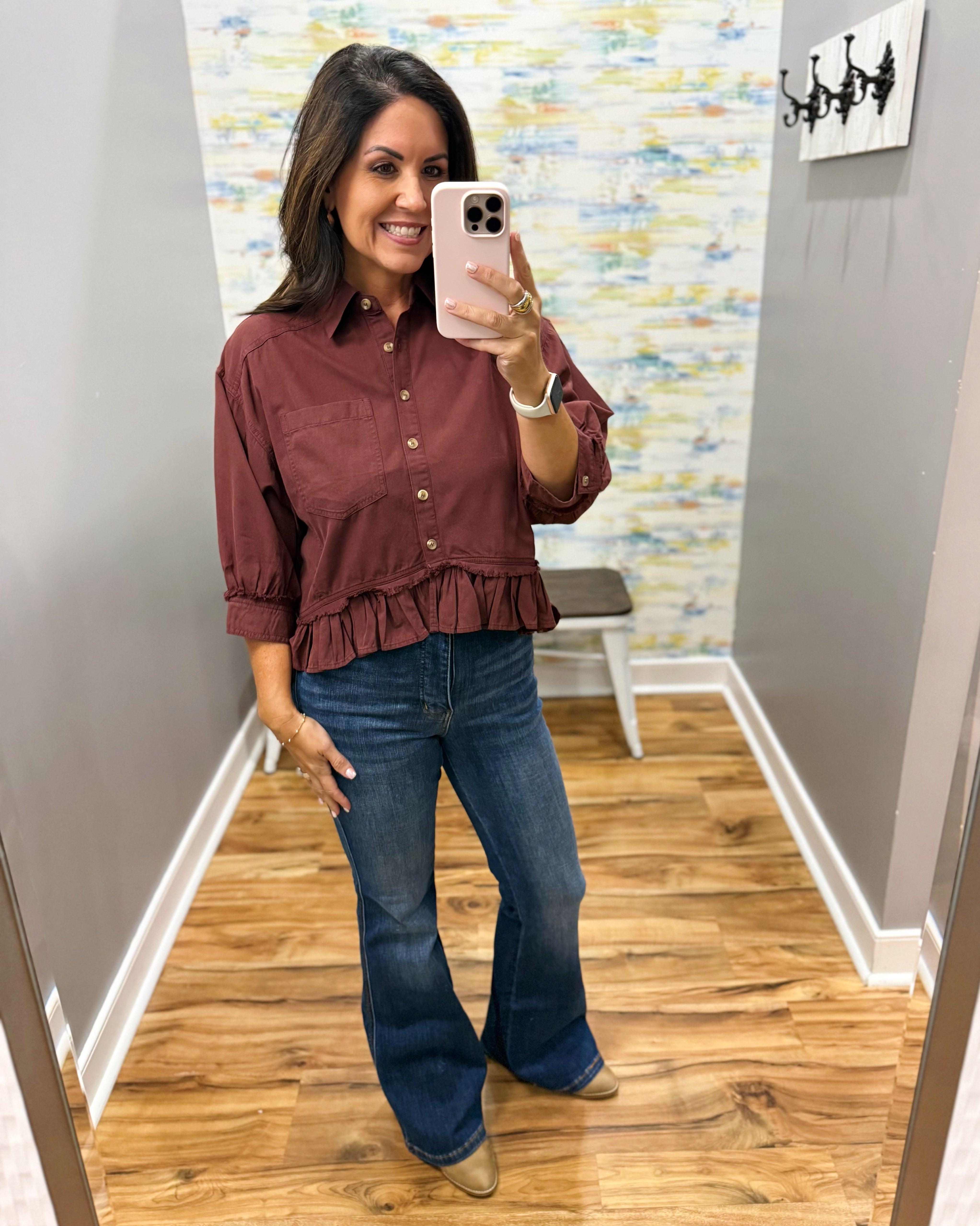 A woman takes a mirror selfie in a fitting room wearing a rust-brown ruffled button-down blouse and blue flare jeans.