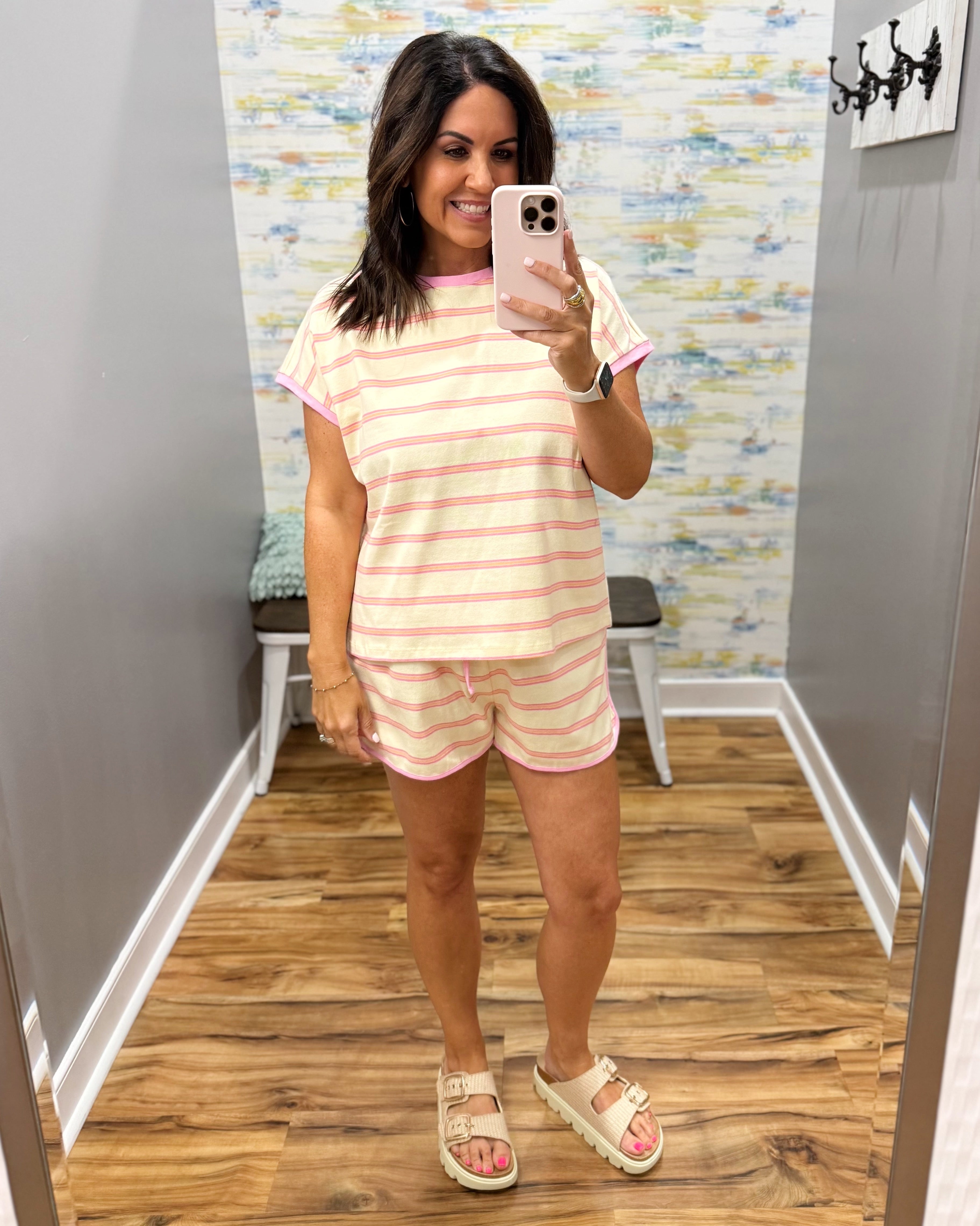 A woman takes a mirror selfie in a fitting room wearing a matching short-sleeve striped pajama set with pink and white horizontal stripes, paired with beige double-strap sandals.