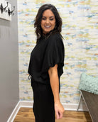 Woman wearing a black outfit standing in a room with patterned wallpaper.