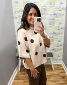 A woman takes a mirror selfie wearing a cream knit sweater with black polka dots and brown pants in a fitting room with wood floors and a patterned wall.