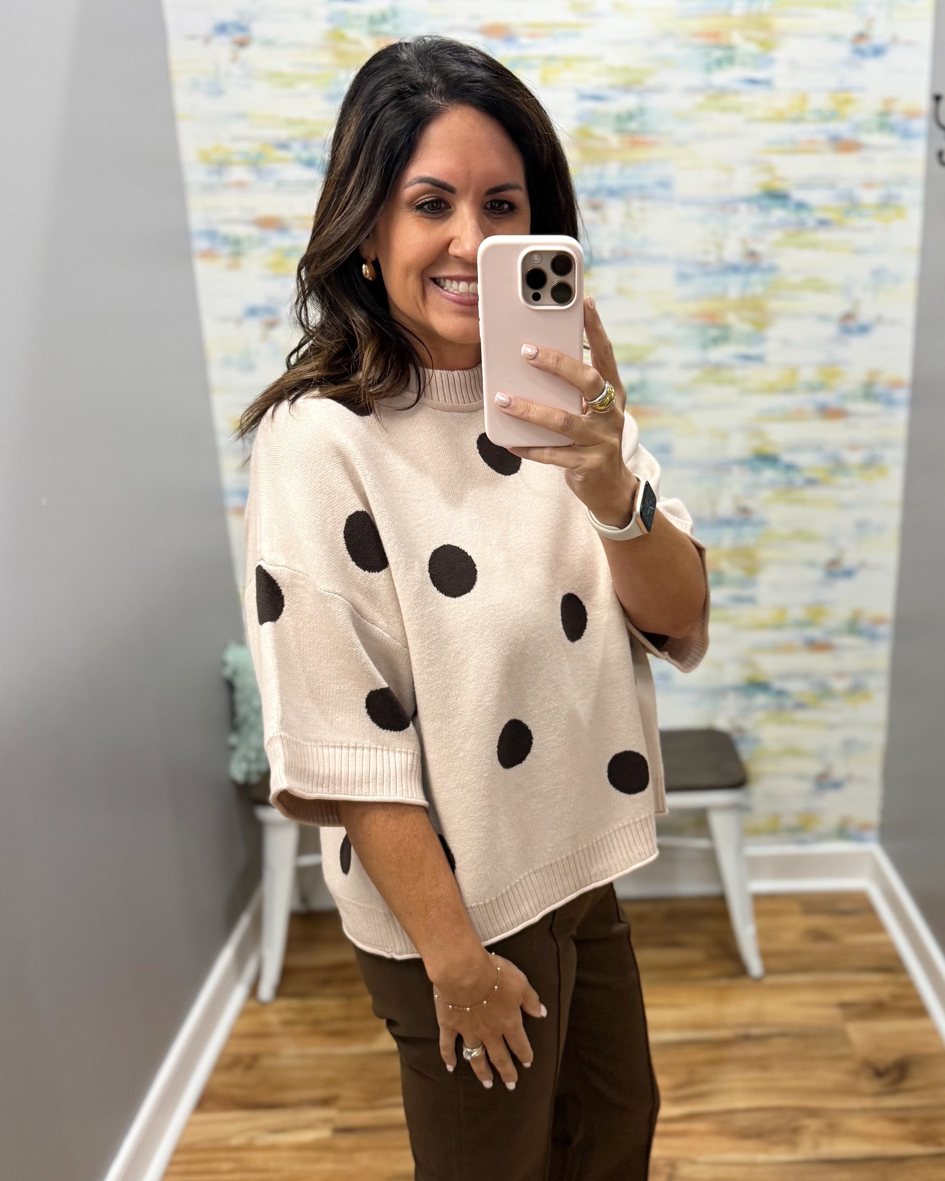 A woman takes a mirror selfie wearing a cream knit sweater with black polka dots and brown pants in a fitting room with wood floors and a patterned wall.