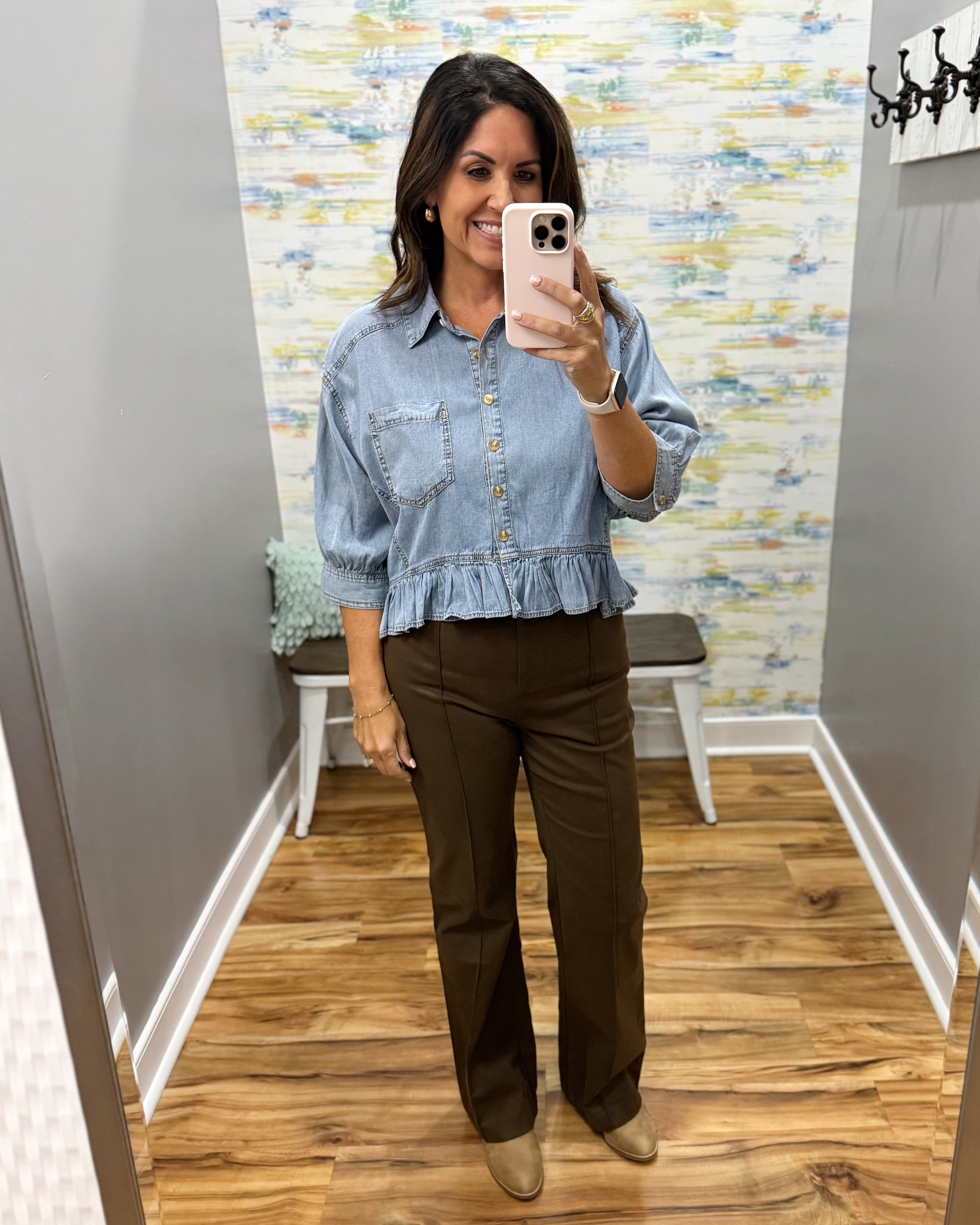A woman takes a mirror selfie in a fitting room wearing a light blue denim shirt with a ruffled hem and brown pants.