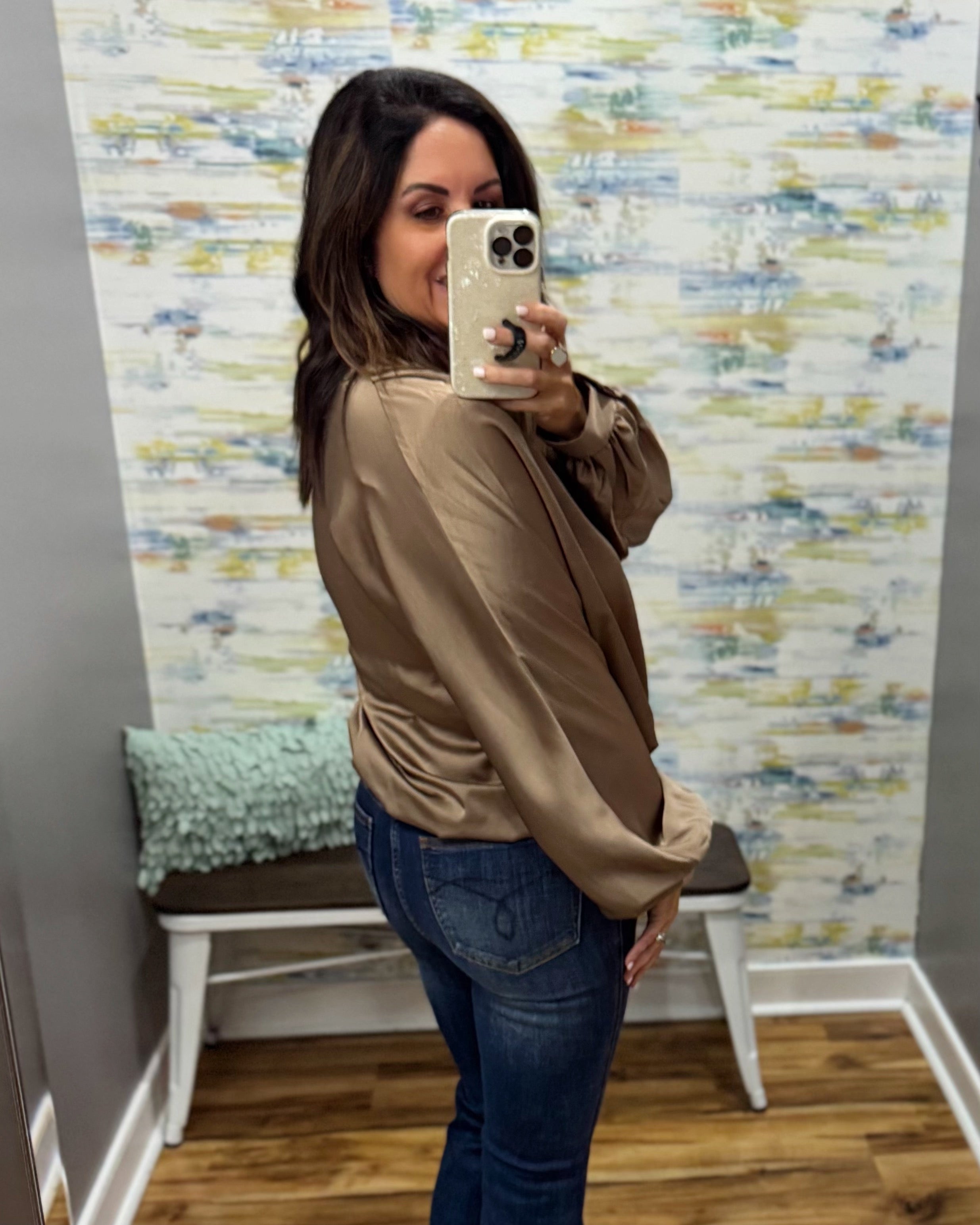 A woman takes a mirror selfie in a fitting room wearing a beige satin long-sleeve blouse and blue jeans. The background features patterned wallpaper, a wooden bench with a green pillow, and wood flooring.