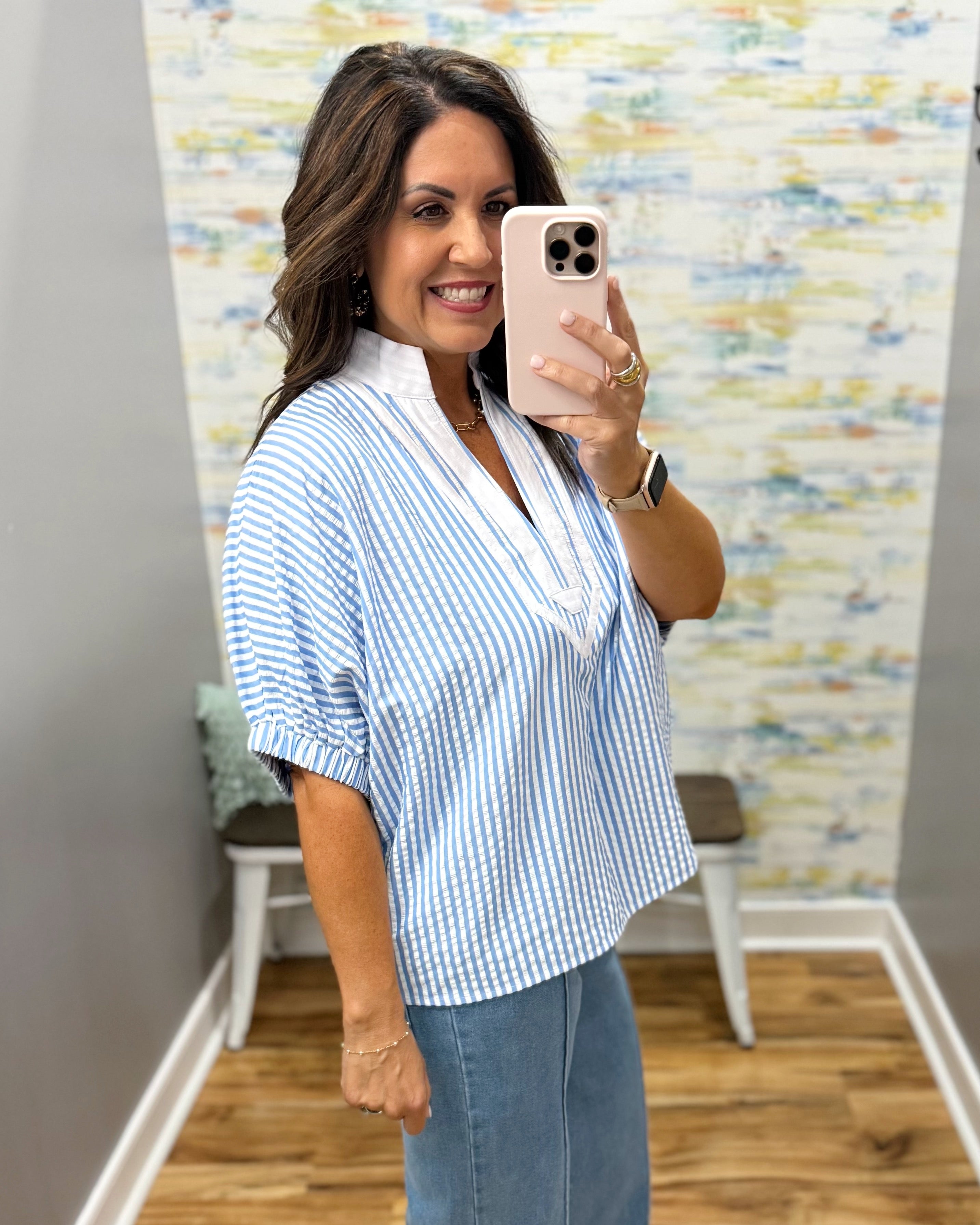 Woman taking a mirror selfie wearing a blue and white striped shirt and jeans, with a colorful wall in the background.