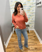 A woman takes a mirror selfie wearing a dusty rose V-neck long-sleeve sweater, light-wash blue jeans, and tan boots in a home setting with wood floors and gray walls.