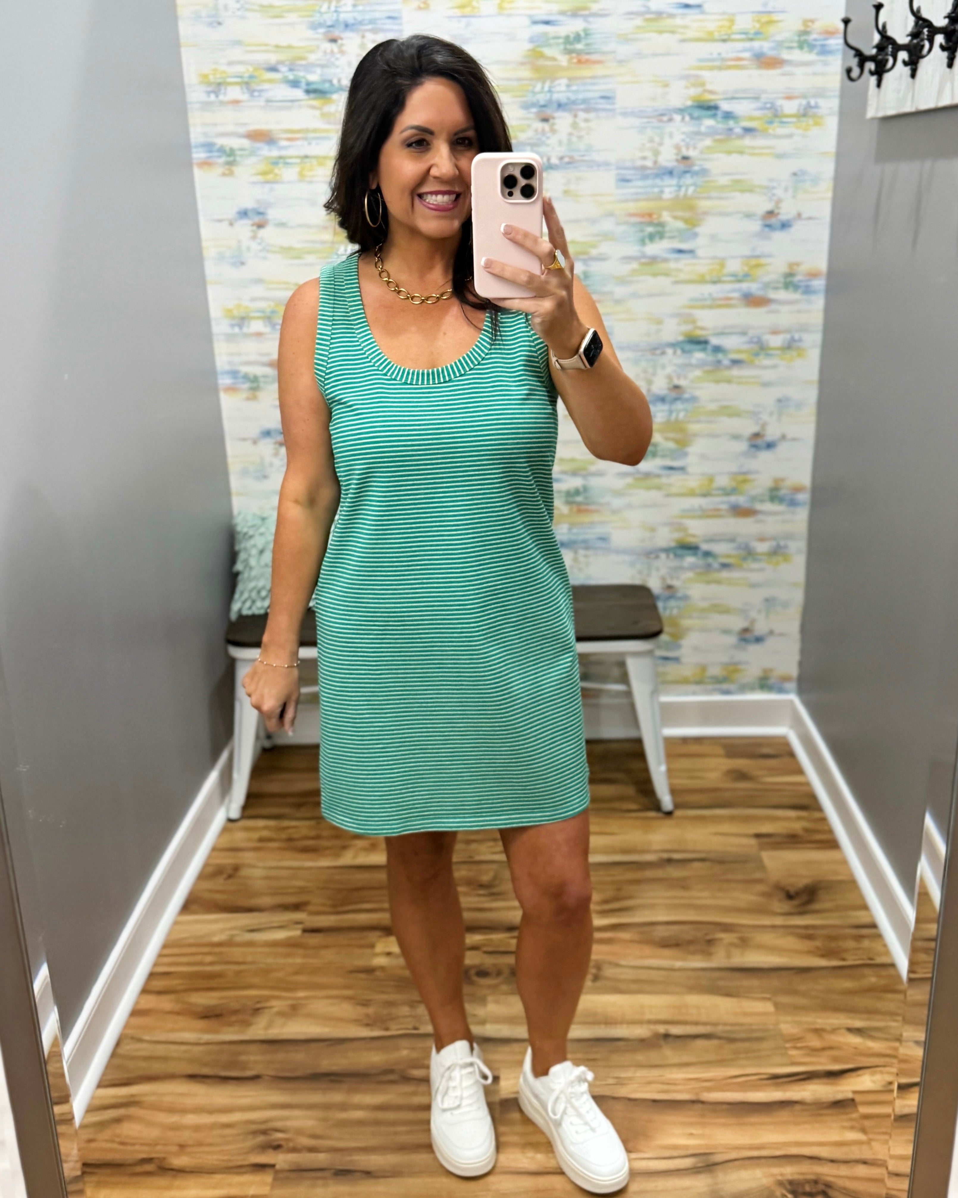 Woman taking a mirror selfie wearing a green dress and white sneakers in a room with patterned wallpaper.