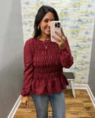 A woman taking a mirror selfie wearing a long-sleeve, smocked, peplum-style top in a deep burgundy red, paired with blue jeans.