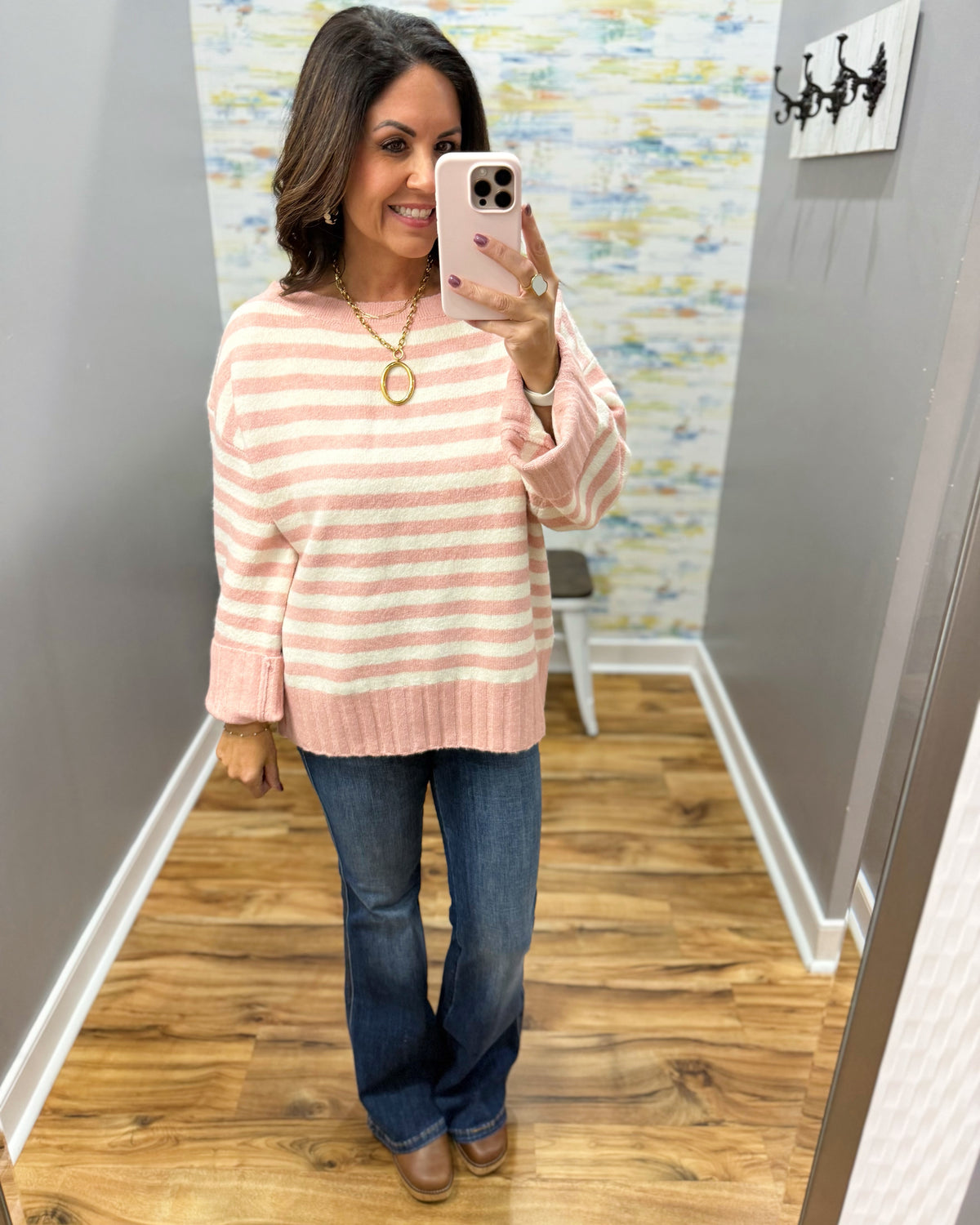 A woman takes a mirror selfie wearing a pink and white horizontally striped knit sweater, blue jeans, and brown boots in a home hallway.