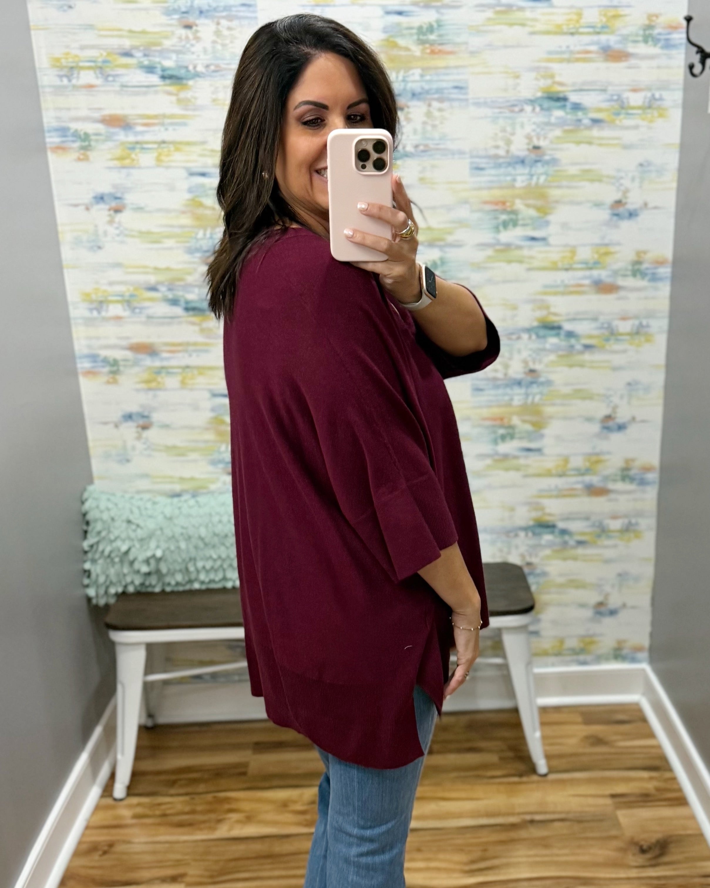 A woman takes a mirror selfie wearing an oversized burgundy top and blue jeans in a room with a patterned wallpaper background and a wooden bench.