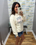 A woman taking a mirror selfie in a fitting room wearing a cream-colored mock-neck sweatshirt with 'SUNNY DAY' printed on the front, paired with denim shorts.