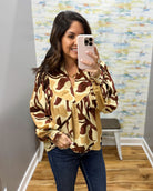 A woman taking a mirror selfie in a fitting room wearing a beige and brown abstract patterned blouse with blue jeans.