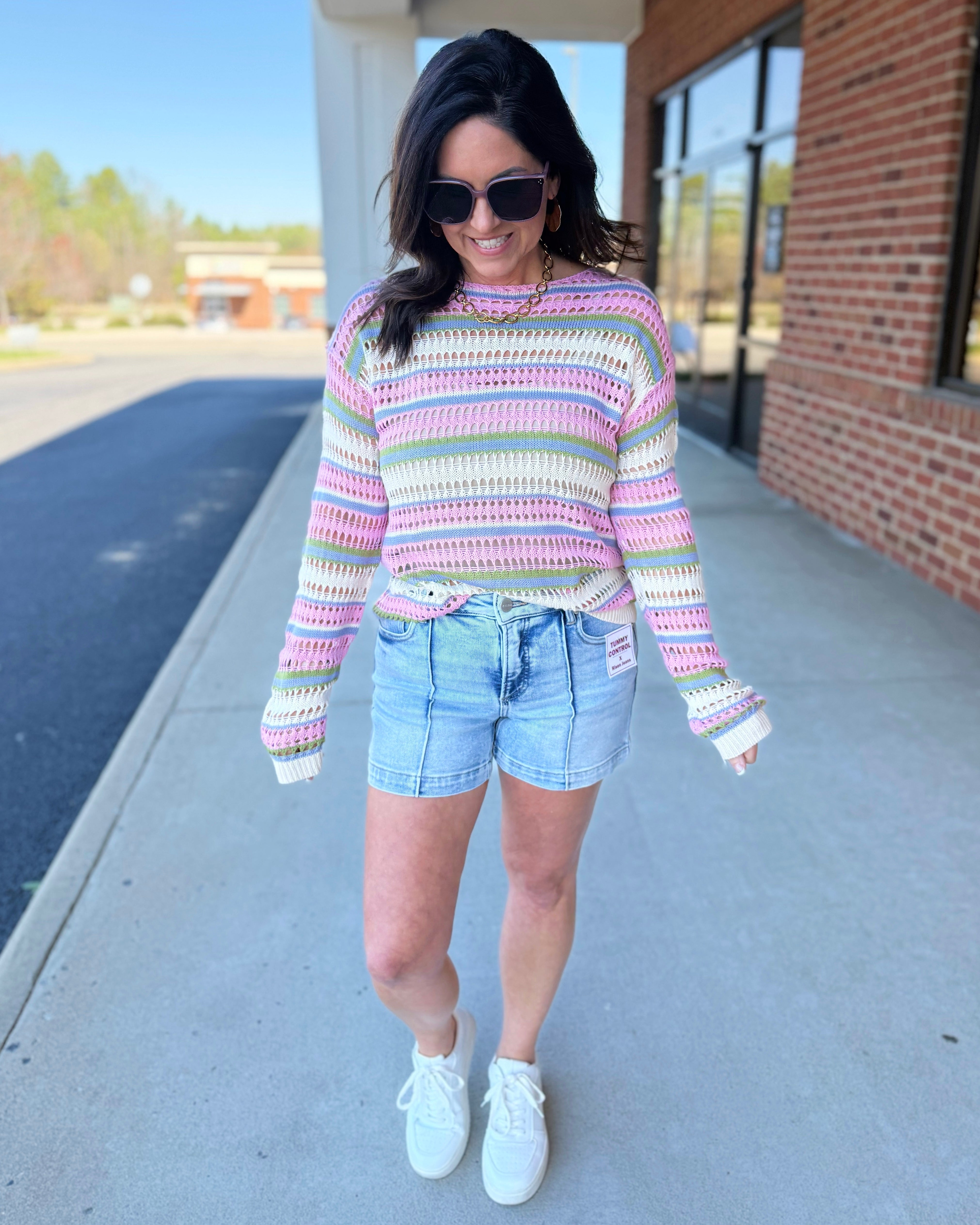 Woman wearing a colorful striped sweater and denim shorts standing on a sidewalk.