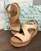 Beige wedge sandals on a wooden surface with a decorative green leafy background